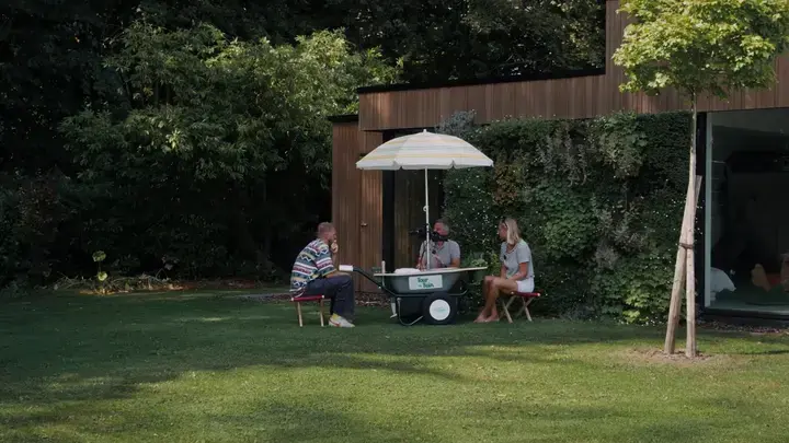 Foto van drie mensen die buiten aan een tafel met parasol zitten in een groene tuin.