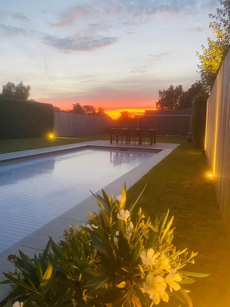 Deze foto toont een prachtig zwembadproject bij zonsondergang. Het zwembad ligt in een moderne tuin met strakke lijnen en sfeervolle verlichting, omgeven door een houten schutting en groen gazon.