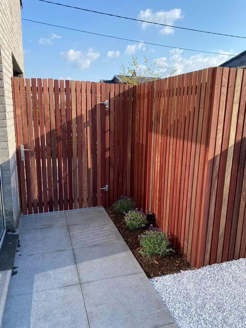 Deze foto toont een moderne en strak afgewerkte achtertuin met een houten schutting. De schutting is gemaakt van verticale houten planken, mooi afgewerkt in een warme houtkleur. In de hoek van de tuin zie je een klein bloembed met paarse bloemen en daarboven zijn er twee metalen sloten op de poort voor beveiliging. De grond is bedekt met grote, lichtgrijze tegels die goed passen bij de rest van de tuin. Rechts van het bloembed ligt een decoratief pad van witte kiezels.