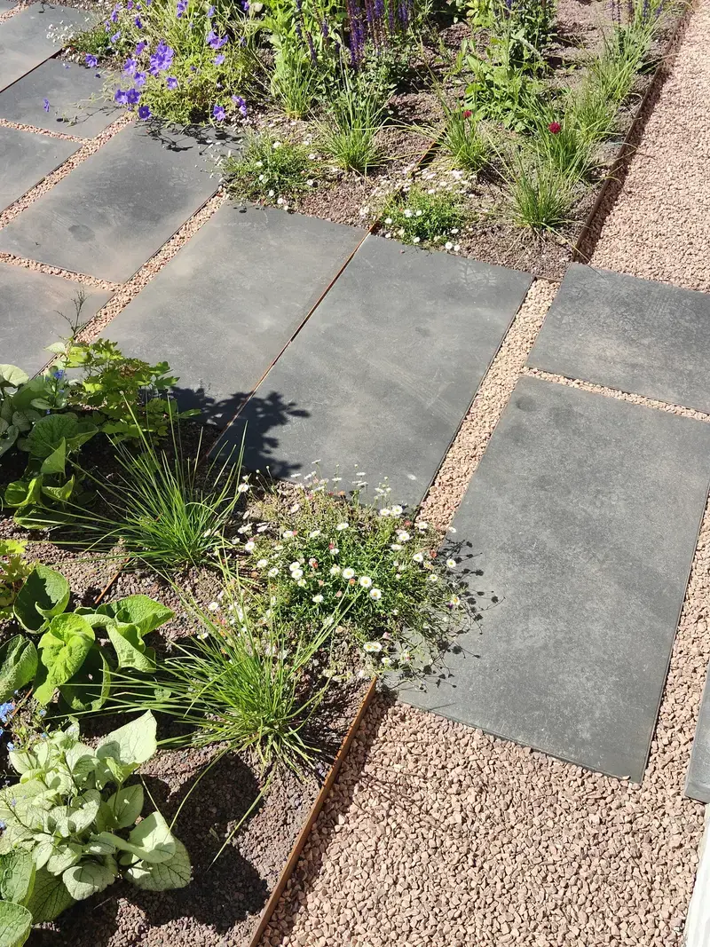 Foto van donkere tuintegels afgewisseld met planten en grindpaden.
