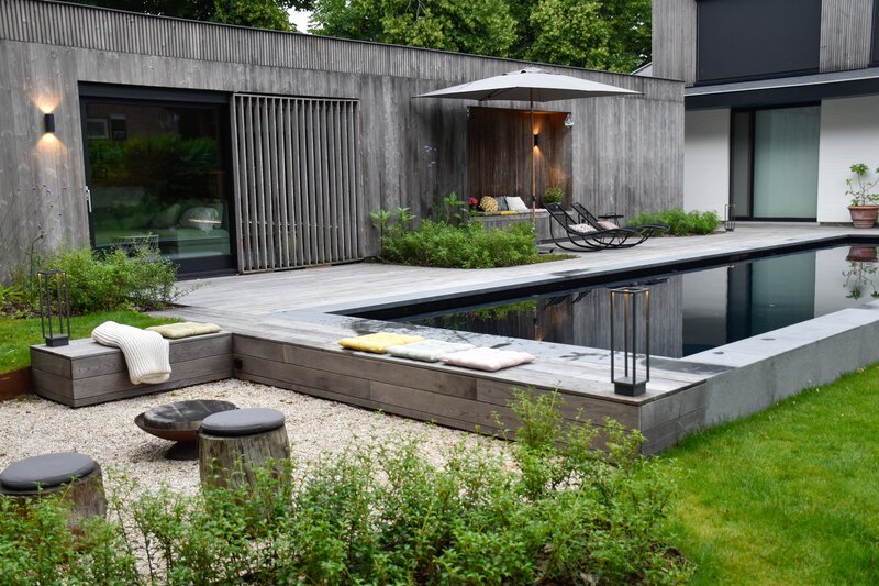 Tuin met natuurlijk ogend zwart zwembad omgeven door houten loungebanken en terrassen om de niveauverschillen op te vangen.