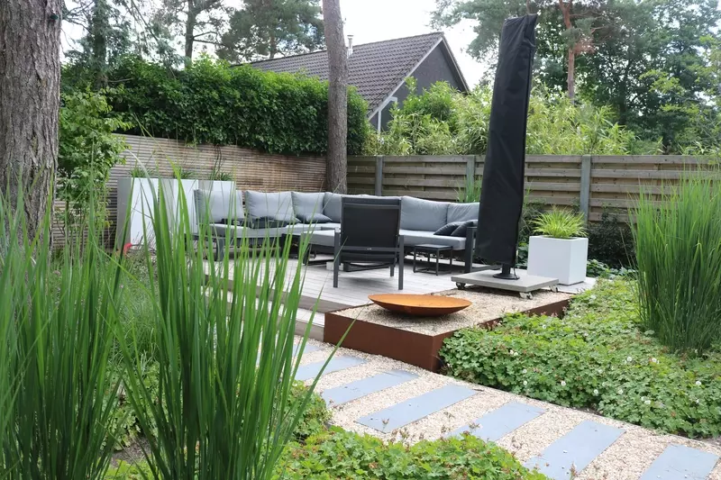 opgaande grassen op de voorgrond. Daarachter een pad met stapstenen die leiden naar een verhoogd houten terras en een vuurkorf in cortenstaal
