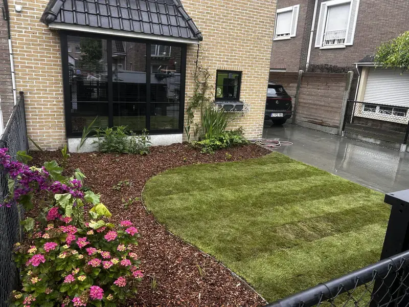 Foto van een kleine voortuin met groen gazon, perkjes met roze bloemen en een beige bakstenen huis.